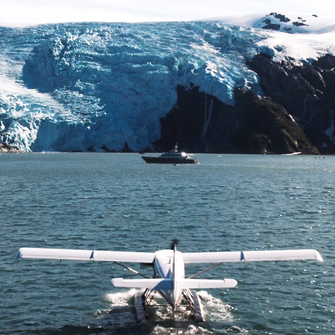 Seaplane landing on. water heading towards a superyacht in front of snow covered mountains
